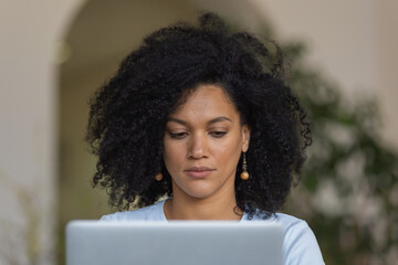 Portrait of a young African American woman typing on laptop keyboard. Brunette with curly hair sitting on yellow sofa in a bright home room. Close up.