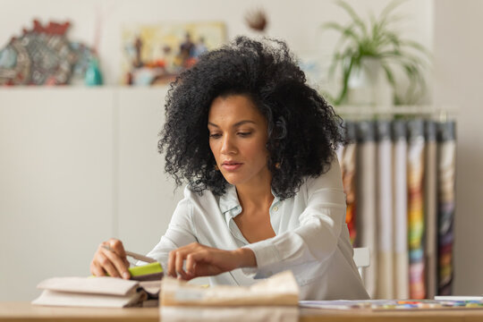 Portrait Of A Young African American Woman Looking Through The Color Palette And Samples Of Fabrics For A Design Project. Female Designer In White Blouse Sits At A Table In Light Office. Close Up.