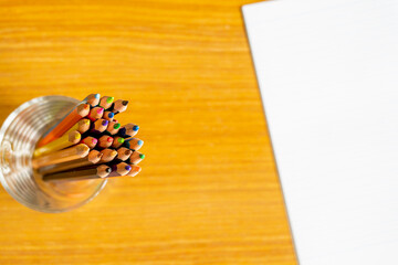 Several colored pencils in glass are placed on a wooden table and a white notebook
