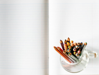 Colored pencils are writing utensils for coloring, arranged in a clear glass on a white notebook