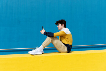 Adolescent sitting on yellow wall using mobile phone against blue wall