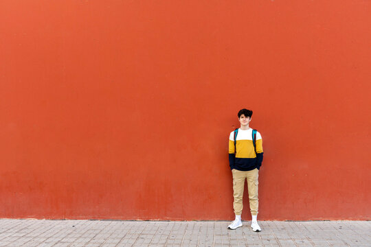 Young male with backpack standing on red wall while looking to camera