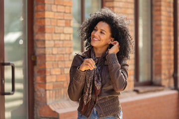 Portrait of young African American woman cute smiling. Brunette with curly hair in leather jacket posing on street against backdrop of blurred brick building. Close up.