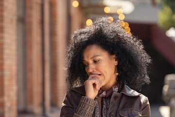 Fototapeta na wymiar Portrait of stylish brooding young African American woman focused thinking about something. Brunette in brown leather jacket posing on street against backdrop of blurred brick building. Close up.