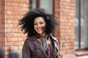 Portrait of stylish young African American woman smiling happily. Brunette with curly hair in brown leather jacket posing on street against backdrop of blurred brick building. Close up.