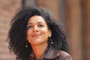 Portrait of stylish brooding young African American woman focused thinking about something. Brunette in brown leather jacket posing on street against backdrop of blurred brick building. Close up.