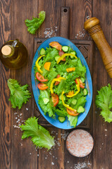 fresh summer green salad with vegetables cucumber, tomato, bell pepper, olive oil, pink salt and leaf lettuce in ceramic bowl, dieting and vegan food on wooden background