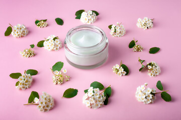 Glass jar with body cream on pink background with small white flowers. Transparent jar with face cream isolated. Cosmetic product for skin care.