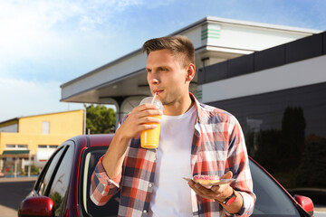 Young man with doughnut drinking juice near car at gas station