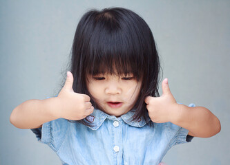 A headshot portrait of a cheerful baby Asian woman, a cute toddler little girl with adorable bangs hair and thumbs up, a child wearing a blue sweater smiling and don't looking to the camera.