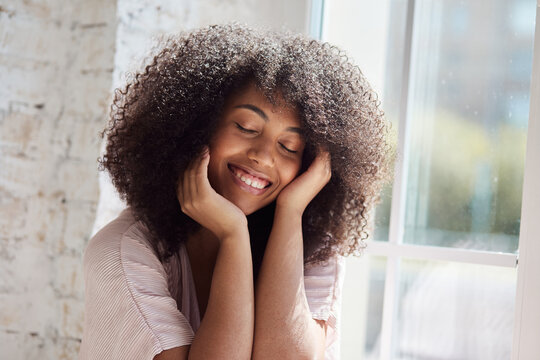 Relaxing smiling young African American woman is sitting near window, resting, and enjoying a lovely stress-free weekend alone at home. In remote work, take a break.