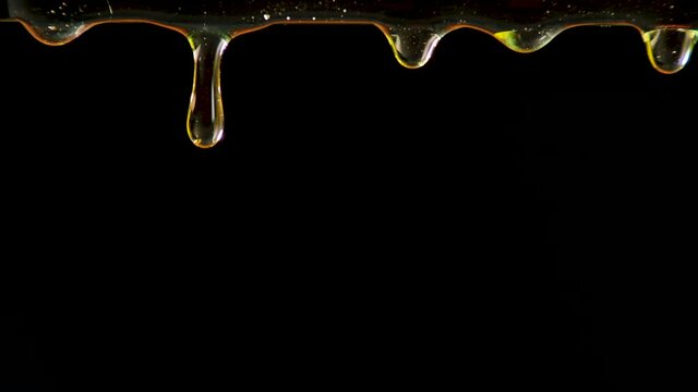 Golden drops of honey drip on a black studio background. The sweet thick liquid drips down. Transparent colorful syrup or oil droplets flowing down. Close up. Slow motion.