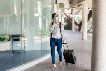 travel, woman in white long-sleeved shirt and jeans, wearing a mask. walking while using mobile phone during travel with suitcase in the left hand and backpack