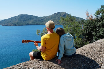 Young man sitting on top of the steep hill and playing a song on the guitar with his girlfriend....