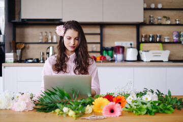 Young brunette woman with long wavy hair working on laptop with flowers on the desk with kitchen in background. Talented florist developing online sales getting ready for workshop.