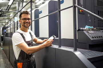 Portrait of graphic engineer standing by modern offset printing machine and checking quality or...