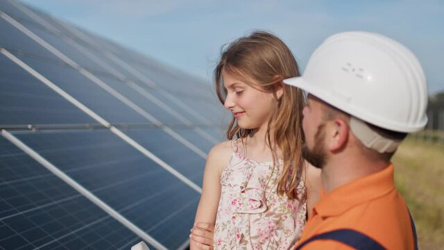 Father with little girl at solar power plant. The father talks about solar energy. The concept of green energy will save the planet for children. The father puts a protective helmet on the girl's head
