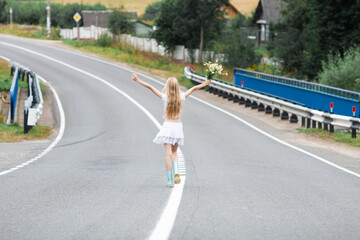 Blonde girl wearing white clothes walking on a paved road with raised arms. Concept of local travel, summer in countryside, finding way.