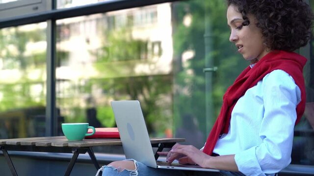 Smiling African American woman using laptop computer, typing on keyboard at workplace. Successful copywriter working online sitting in cafe