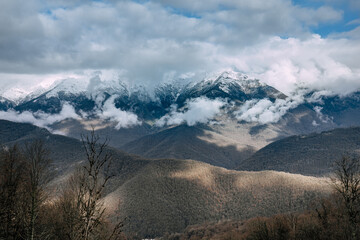 Beautiful view to snow-capped mountains above dense cloud in sunshine. Scenic mountain landscape with white-snow peaks among dense low clouds. Krasnaya Polyana,Caucasus Mountains. Mist swirling around