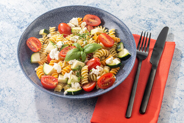 Italian pasta fusilli tricolore with cherry tomatoes, zucchini and feta cheese in a blue shallow bowl on a red napkin with black silverware on a blue surface