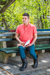 Dressing in a red Polo shirt, green pants and long leather boots, a young handsome guy is sitting on a long chair  in the morning of a spring season, reading a book.