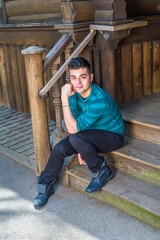 Dressing in a green striped long sleeves Henley shirt, black pants and leather boots, a young handsome guy is sitting on stairs outside a wooden house and taking a break..