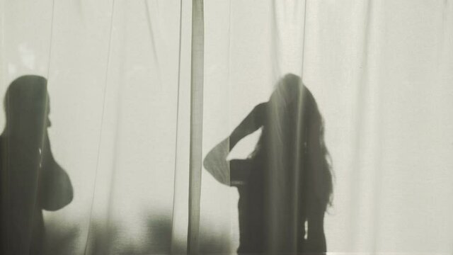 Silhouette of a long-haired woman and bold man standing behind curtains with their smartphones. Contrasting shadow in the bright sun. Summer mood. Young couple on vacation. People talking phone. Call.