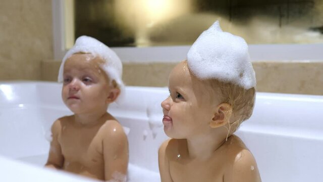 Adorable tired white twins taking bath with soap bubbles on their heads getting blown off. Happy childhood concept