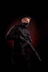 A man in dark clothes holds an automatic carbine in his hands. The face is covered with a mask, hands are in gloves. A figure with a weapon on a dark back with a red light spot.