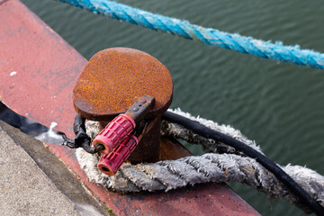 Ein Seil und ein Elektrokabel mit Klemmen an einem Poller im Hafen.