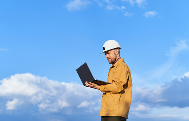 Attractive caucasian bearded supervisor, building contractor or engineer in white helmet and orange jacket using laptop at construction site. Sunny day with blue cloudy sky. High quality image