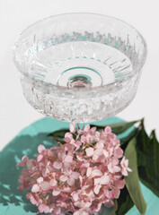 beautiful crystal high glass with mineral water on a turquoise table next to pink hydrangea flowers. water drinking and wellness concept. vertical size.