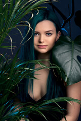 Girl with blue green hair on a gray background, blue backlighting. Tropical plants around. Bright colored appearance of a creative person