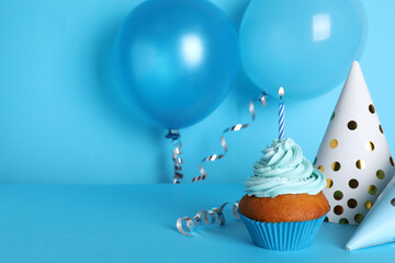 Delicious birthday cupcake with candle near party hats and balloons on light blue background, space...