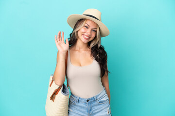 Fototapeta premium Teenager Russian girl with pamel and beach bag isolated on blue background saluting with hand with happy expression