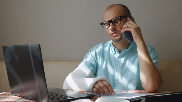 Man in eyeglasses with arm in plaster cast talking on phone and using laptop, sunshine through window blinds on his blue shirt. Working remotely from home after accident. Concept of online business