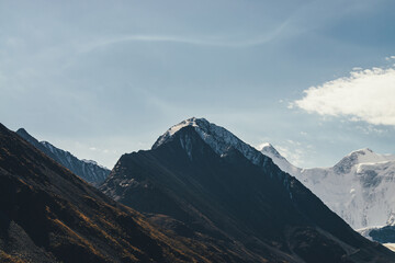 Atmospheric alpine landscape with high mountain silhouette with snow on peaked top under cirrus clouds. Dramatic mountain scenery with beautiful snow-covered pointy peak and high snowy mountain wall.