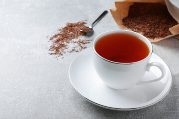 Ceramic cup of aromatic rooibos tea, spoon and scattered dry leaves on light grey table. Space for...