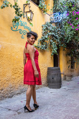 Mid adult afro mexican smiling woman wearing a red dress and black high heels walking an alley