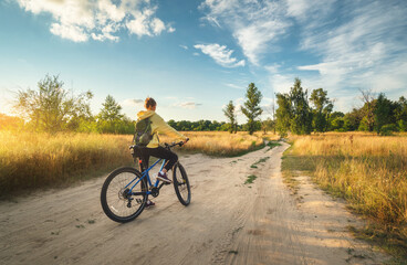 Fototapeta na wymiar Woman is riding a mountain bike in cross country road at sunset in summer. Colorful landscape with sporty girl with backpack riding bicycle, field, dirt road, green grass, blue sky. Sport and travel