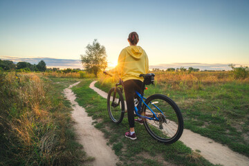 Obraz premium Woman is riding a mountain bike in cross country road at sunset in summer. Colorful landscape with sporty girl in yellow hoodie, bicycle, field, dirt road, green grass, trees, sky. Sport and travel