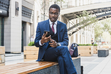 Portrait of a black African American businessman in a suit, glasses and headphones sits on a bench...