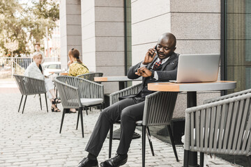 Portrait of a black African American businessman in a suit sitting in a city cafe outdoors and talking on the phone.