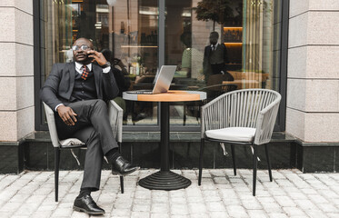 Portrait of a black African American businessman in a suit sitting in a city cafe outdoors and talking on the phone.