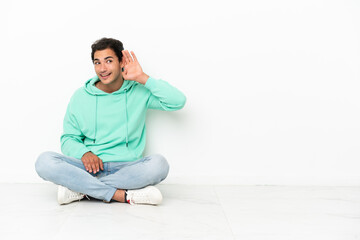 Caucasian handsome man sitting on the floor listening to something by putting hand on the ear