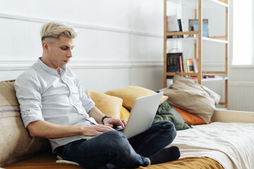 Young attractive smiling blonde guy is browsing at his laptop, sitting at home on the cozy sofa at home, wearing casual outfit