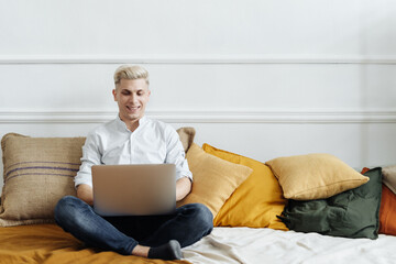 Young attractive smiling blonde guy is browsing at his laptop, sitting at home on the cozy sofa at home, wearing casual outfit