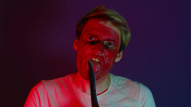 Angry man, maniac or crazy, licks a knife standing in a dark room. Portrait man with a knife. Concept: Serial killer maniac.