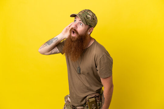 Redhead Military man with dog tag isolated on yellow background listening to something by putting hand on the ear